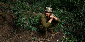 Ein erschöpfter Soldat sitzt allein an einem Abhang und raucht eine Zigarette, er trägt Kampfkleidung und einen kleinen Hut