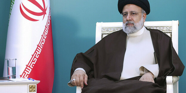 Ebrahim Raisi sitzt auf einem weißen Stuhl, neben ihm aufgestellt eine iranische Fahne