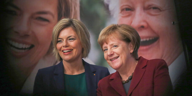 Julia Klöckner und Angela Merkel stehen lächelnd vor einem Plakat mit lächelnden Gesichtern.