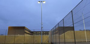 ein hoher Zaun, helle Beleuchtung und ein Gefängnisgebäude