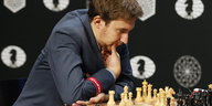 Ein Mann in einem blauen Jacket sitzt vor einem Schachbrett, eine Hand hat er am Kinn