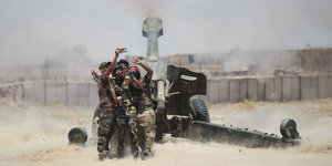 vier Männer in Militärkleidung machen ein Selfie vor einem noch rauchenden Artilleriegeschoss