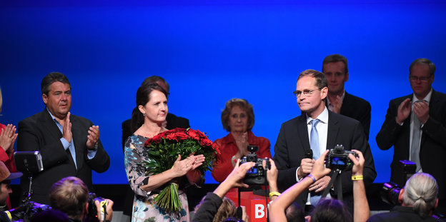 Mehrere Menschen, darunter eine Frau mit einem großen roten Blumenstrauß stehen auf einer Bühne vor einem blauen Hintergrund