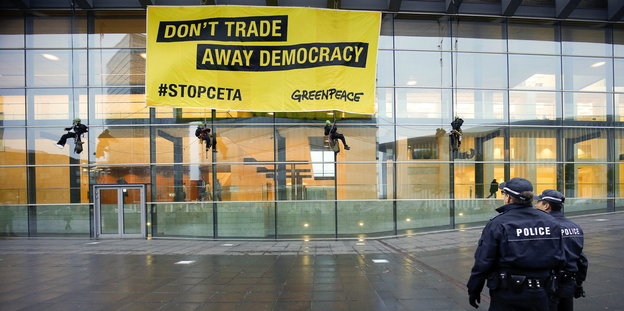 An einem Gebäude hängt ein Plakat „Don't tade away democracy #Stopceta Greenpeace“, zwei Polizisten blicken darauf
