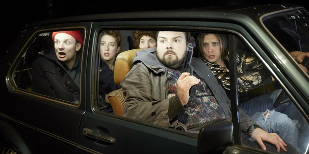 Vier Schauspieler sitzen in einem Auto, einer hat eine Pistole in der Hand