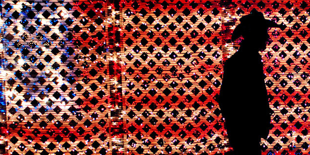 Die dunkle Silhouette eines Mannes vor einer perforierten Stars-and-Stripes-Flagge Die dunkle Silhouette eines Mannes vor einer perforierten Stars-and-Stripes-Flagge