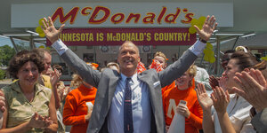 Ein Mann in Anzug steht inmitten einer Menschengruppe vor einem McDonalds-Restaurant und hält die Arme hoch