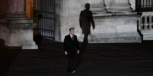 Emmanuel Macrons kommt im Dunkeln aus einem Gebäude, sein Schatten überragt ihn riesig an dessen steinerner Wand