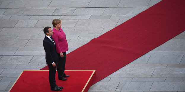 Macron steht im schwarzen Anzug neben Merkel auf einem roten Podest Macron steht im schwarzen Anzug neben Merkel auf einem roten Podest