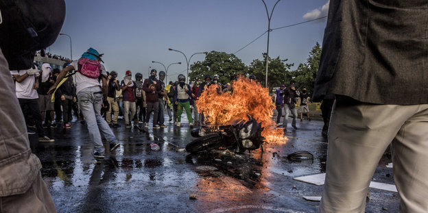 Ein brennendes Mottorrad liegt auf einer Straße, darum stehen mehrere Menschen