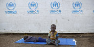 Auf einer blauen Decke liegt ein kleiner Junge und schläft, neben ihm sitzt sein kleiner Bruder, auf der Wand hinter ihnen ist das Logo des UN-Flüchtlingshilfswerks gemalt