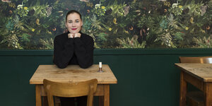 Eine Frau sitzt an einem Holztisch, hinter ihr eine Tapete mit Pflanzenmuster