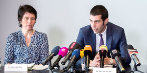 Eine Frau und ein Mann sitzen an einem Tisch auf dem viele Mikrophone stehen.
