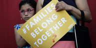 Ein Mädchen steht neben ihrer Mutter, die ein Schild mit der Aufschrift „Families belong together“ – „Familien gehören zusammen“