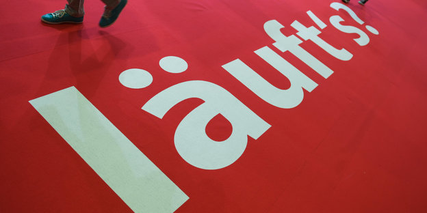 Besucher der Frankfurter Buchmesse gehen in einer Halle über einen Teppichboden, auf dem des Wort "läuft's ?" gedruckt ist Besucher der Frankfurter Buchmesse gehen in einer Halle über einen Teppichboden, auf dem des Wort "läuft's ?" gedruckt ist