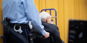 Ein Justizbeamter schiebt den Angeklagten, er sitzt im Rollstuhl, in den Gerichtssaal