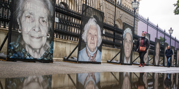 An einem Zaun in einer nassen Straße hängen große Portraits älterer Menschen.