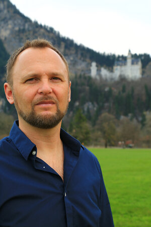 Markus Richter trägt ein blaues Hemd. im Hintergrund ist das Schloss Neuschwanstein zu sehen.
