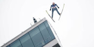 ein Skispringer fliegt auf die Kamera zu. Im Hintergrund die Fensterfront der Schanze