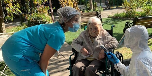 Eine alte Frau in einem Rollstuhl mit zwei Pflegerinnen.