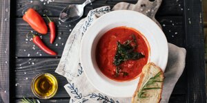 Ein Teller mit roter Suppe mit Brot serviert, auf dem Tisch darum sind Chilis, ein Glas OIlvenöl, ein Löffel