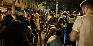 Dicht gedrängt stehen und sitzen junge Männer und Frauen nachts im Licht einer Laterne an der Hamburger Schanze. Einige Polizist:innen in Uniform und Mundschutz stehen dabei