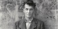 Ludwig Wittgenstein steht vor einer grauen Wand auf der Kratzer und Botschaften eingeritzt sind