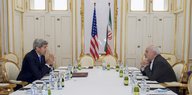 zwei Männer sitzen sich an einem gedeckten Tisch gegenüber, dahinter die Fahnen von USA und Iran