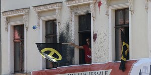 Ein Mann schwingt eine Fahne mit dem Logo der Identitären Bewegung aus einem Fenster. Eine Frau schaut in die Kamera