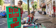 Zu sehen sind zwei Aktivistinnen von Klimaneustart Berlin. Sie tragen beide eine rote Weste. Eine der beiden befestigt gerade ein grünes Plakat mit der Aufschrift "Es ist nicht zu spät" an einem Baum. Die andere Aktivisten hält mehr Plakate fest, die auf