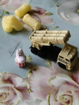 Auf einem Tisch mit Tischdecke steht ein Spielzeug-Panzerabwehrwagen, eine Moomin-Mama und ein paar Frühstücksflocken