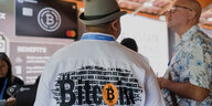 Ein Mann steht auf einer Messe mit der Aufschrift "Bitcoin" auf seinem Hemd