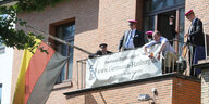 Mitglieder der Burschenschaft Germania stehen und sitzen auf einem Balkon, trinken Bier und beobachten eine Demonstration vor ihrem Haus.