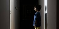 Ein kleiner Junge steht frontal vor einer offenen Tür in einem Hausflur und wird vom herausscheinenden Licht angestrahlt