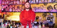 Ein Mann mit rotem Shirt und verschränkten Armen steht hinter einer Bar, an die Wand sind Quallen gemalt, die zwischen Flaschen schweben