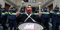 Eine Fru mit Trommel und einem roten Klebeband über dem Mund steht vor einer Reihe Polizisten