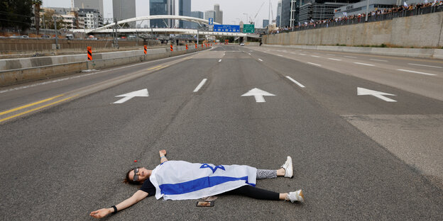 Eine Frau liegt rücklings mitten auf einer leeren Autobahn. Sie hat die israelische Flagge mit dem blauen Davidstern über sich ausgebreitet
