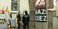 Zwei Polizisten stehen im Hintergrund, im Vordergrund eine Säule mit dem Aufkleber "Nazi Kiez"