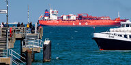 Ein rotes Schiff mit der Aufschrift "LNG" vor der Küste von Binz. Davor ein Passagierboot mit der Aufschrift "Binz". Am Ufer stehen mehrere Menschen auf einem Steg.