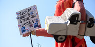 Ein Mensch trägt ein Pappmaché-Auto und eine Schild, auf dem steht: "Rechte nichts steuern lassen"