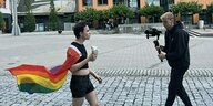 Eine junge Person in Shorts mit Regenbogenfahne isst einen Döner und wird von einer anderen Person gefilmt