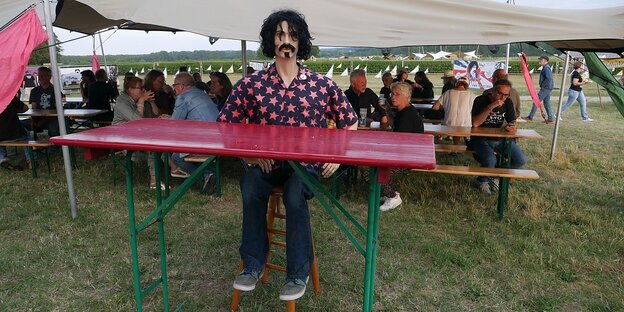 Eine Puppe, die Aussieht wie Frank Zappa, sitzt auf einer Bierbank