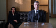 Das Standphoto zeigt Oppenheimer in anzug und Krawatte, irritiert blickend. Schräg hinter ihm seine Frau auf einer Couch