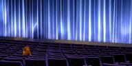 Eine einzelne Kinobesucherin sitzt in einem leeren Kinosaal, die Leinwand ist von einem blauen Vorhang verhängt
