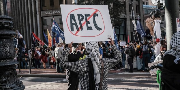 Eine Person mit PaliTuch um den Kopf hält ein Schild hoch, auf dem die Aufschrift APEC durchgestrichen ist