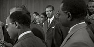 Patrice Lumumba steht inmitten einer Gruppe Männer in einem Raum.