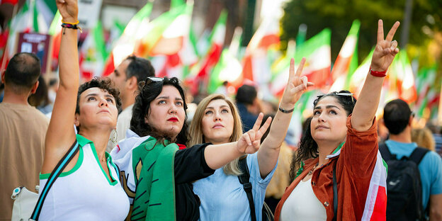 Frauen stehen zusammen, schauen Richtung Himmel und formen mit zwei Fingern das Victory-Zeichen, im Hintergrund eine Menschenmenge mit iranischen Fahnen