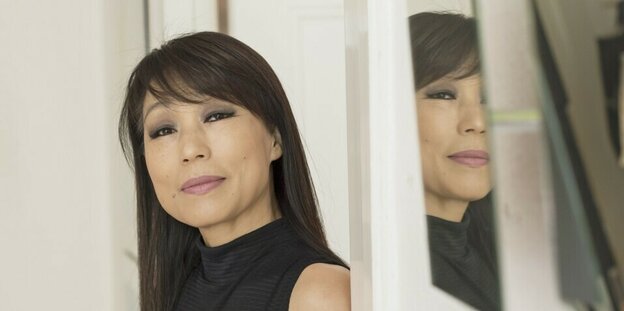 Porträtaufnahme von Unsuk Chin. Die Musikerin ist in der Halbfigur zu sehen. Sie trägt ein schwarzes Kleid. An der Wand neben ihr spiegelt sich ihr Gesicht in einem Stück Spiegelglas.