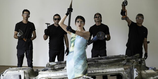 Die Künstlerin steht in bläulichem Kleid vor Schrottteilen. In ihrer erhobenen Hand hält sie eine Axt. Vier schwarzgekleidete Männer im Hintergrund halten Hammer und Flex in die Höhe.