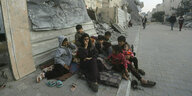 Eine FRau und Kinder sitzen vor einem zerstörten Haus auf dem Straßenpflaster